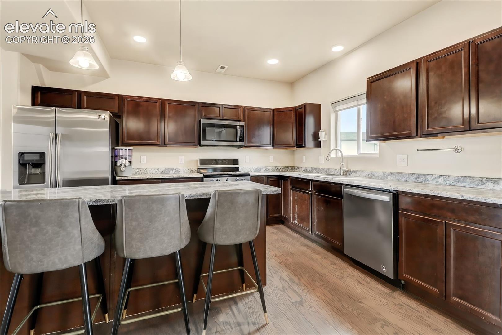 Kitchen featuring dark wood cabinetry, stone-surface countertops, wood-finish flooring, stainless steel appliances, and dual pendant lighting