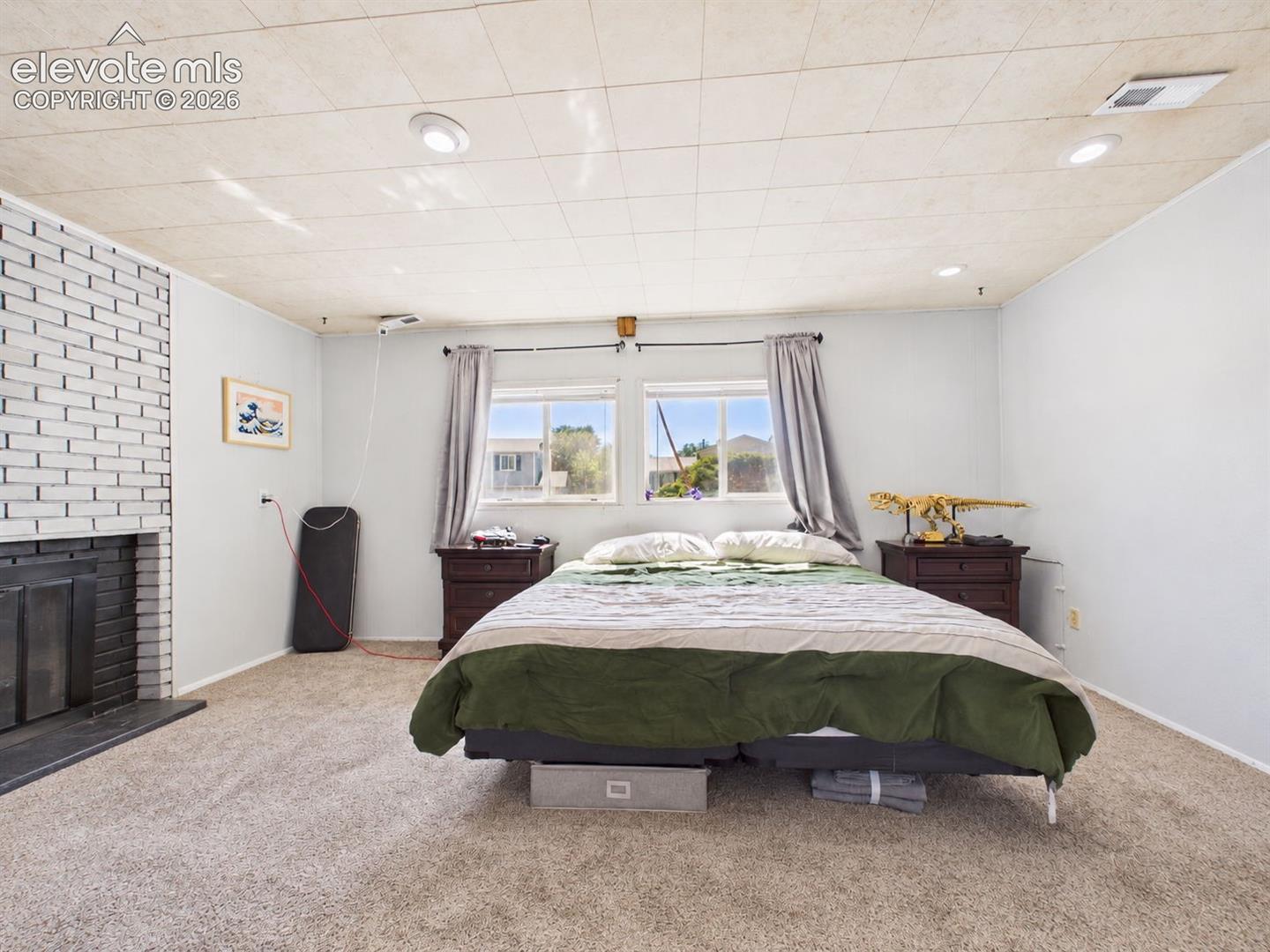 Spacious bedroom features a distinctive painted-brick fireplace, recessed lighting, carpeting, and dual windows that bring in abundant natural light.