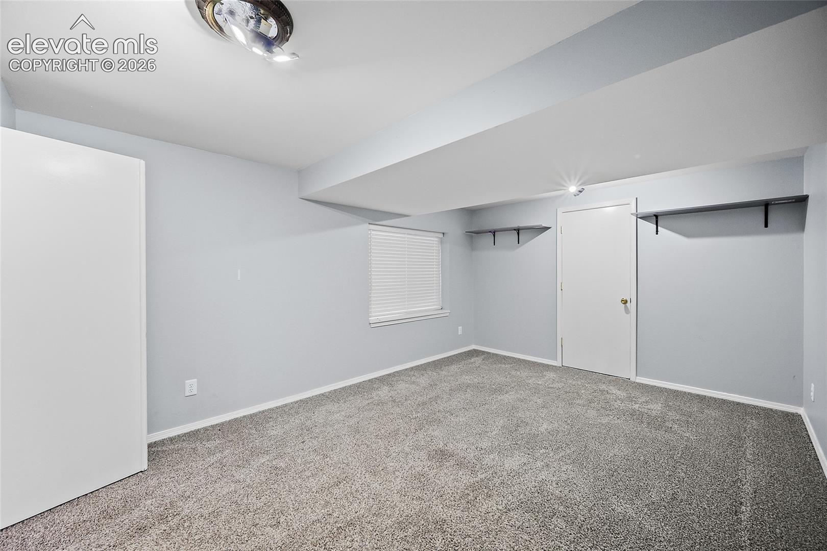 Plush gray carpet, recessed ceiling beam, window with blinds, white door, and track lighting