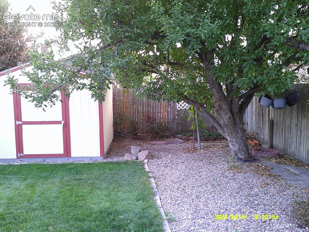 Expansive backyard featuring a manicured lawn, established shade tree, gravel landscaping, a privacy fence, and an exterior storage shed