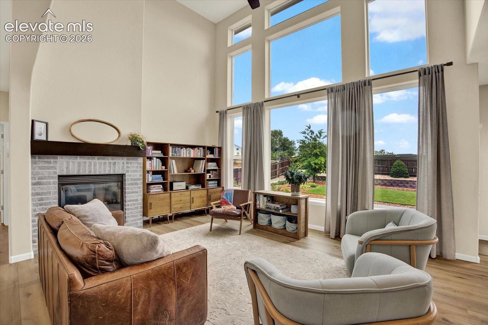 Double-height ceilings, floor-to-ceiling windows, brick fireplace with a wood mantel, and wood-finish flooring