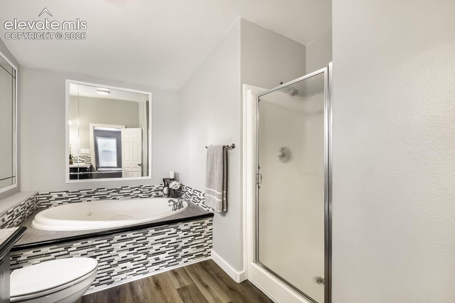 Jetted soaking tub with decorative mosaic tile surround, walk-in shower with metal-framed glass door, wood-finish flooring, and large wall-mounted mirror
