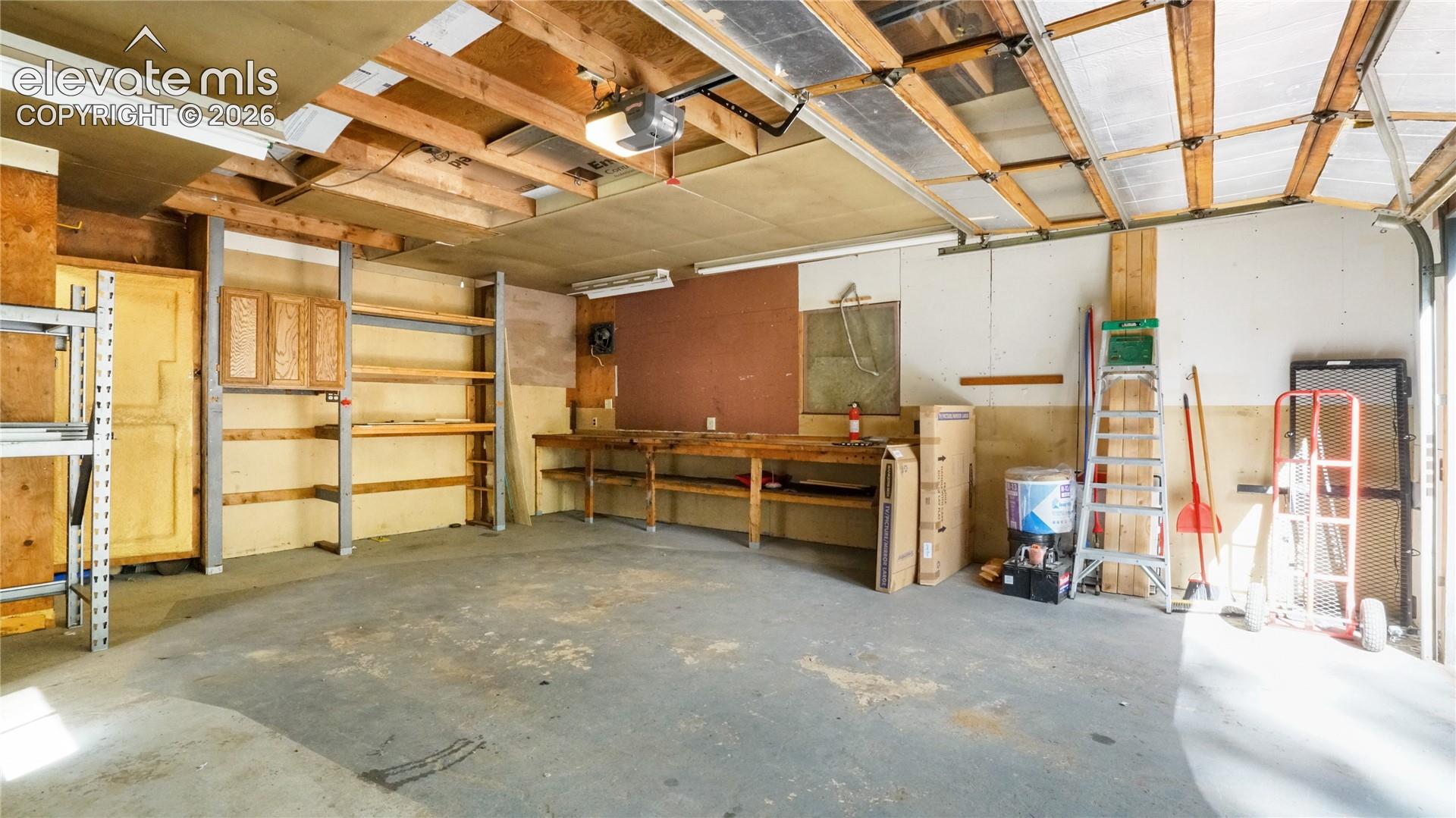 Concrete flooring with exposed ceiling joists, built-in shelving, wood cabinetry, and an overhead garage door
