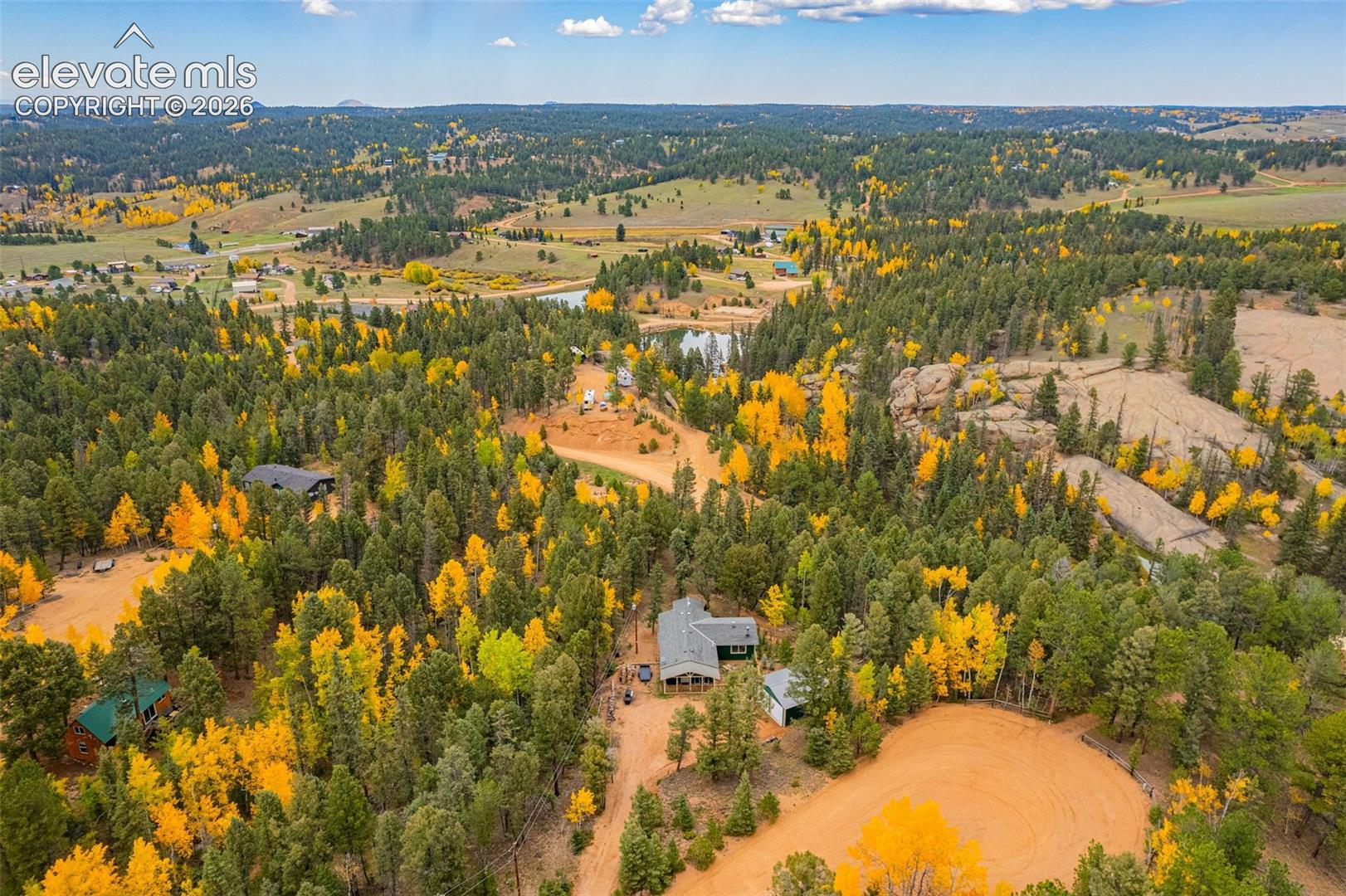 Aerial perspective of a multi-structure property featuring dense evergreen and deciduous forest canopy, natural rock formations, and a private earthen access drive