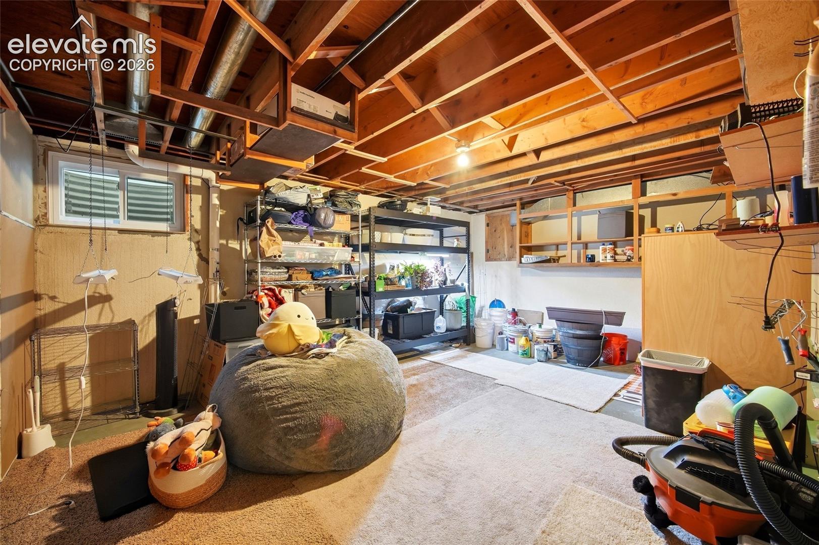 Unfinished lower level featuring exposed wood-joist ceiling, partial wall shelving, and an egress window