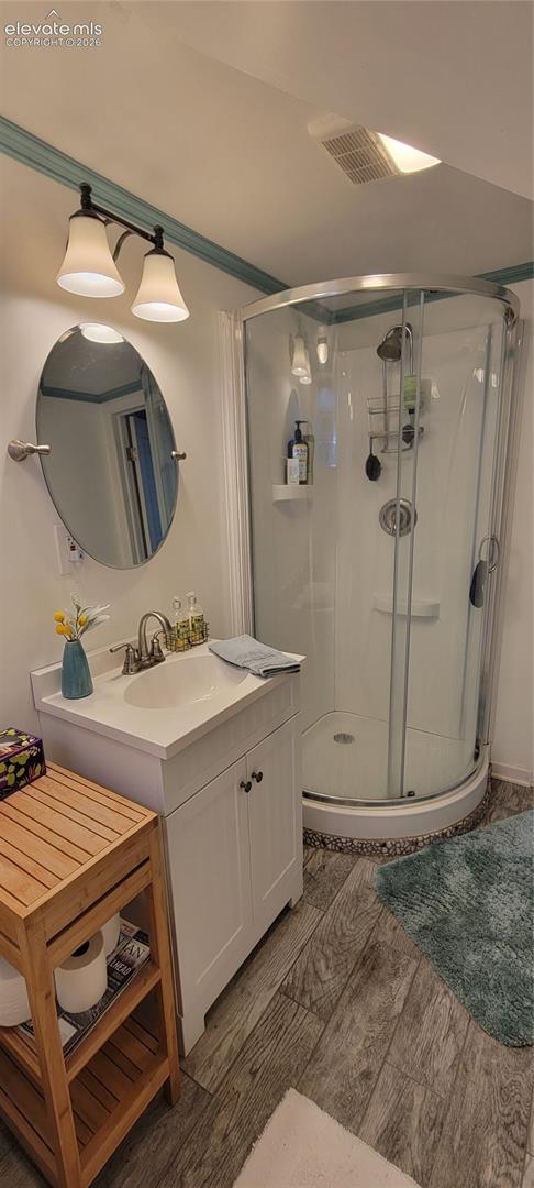 Downstairs bathroom featuring a glass-enclosed corner shower, white vanity with integrated sink, oval wall mirror, modern two-light fixture, and wood-finish flooring