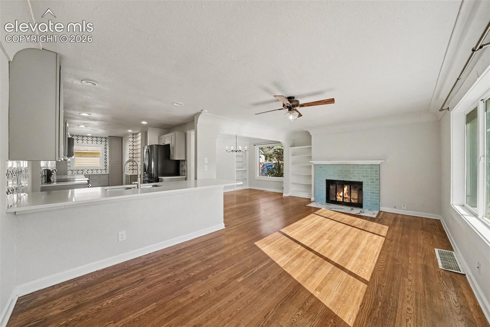 Open concept kitchen featuring a peninsula with countertop seating, wood-finish flooring, a brick fireplace with a painted hearth, and built-in shelving