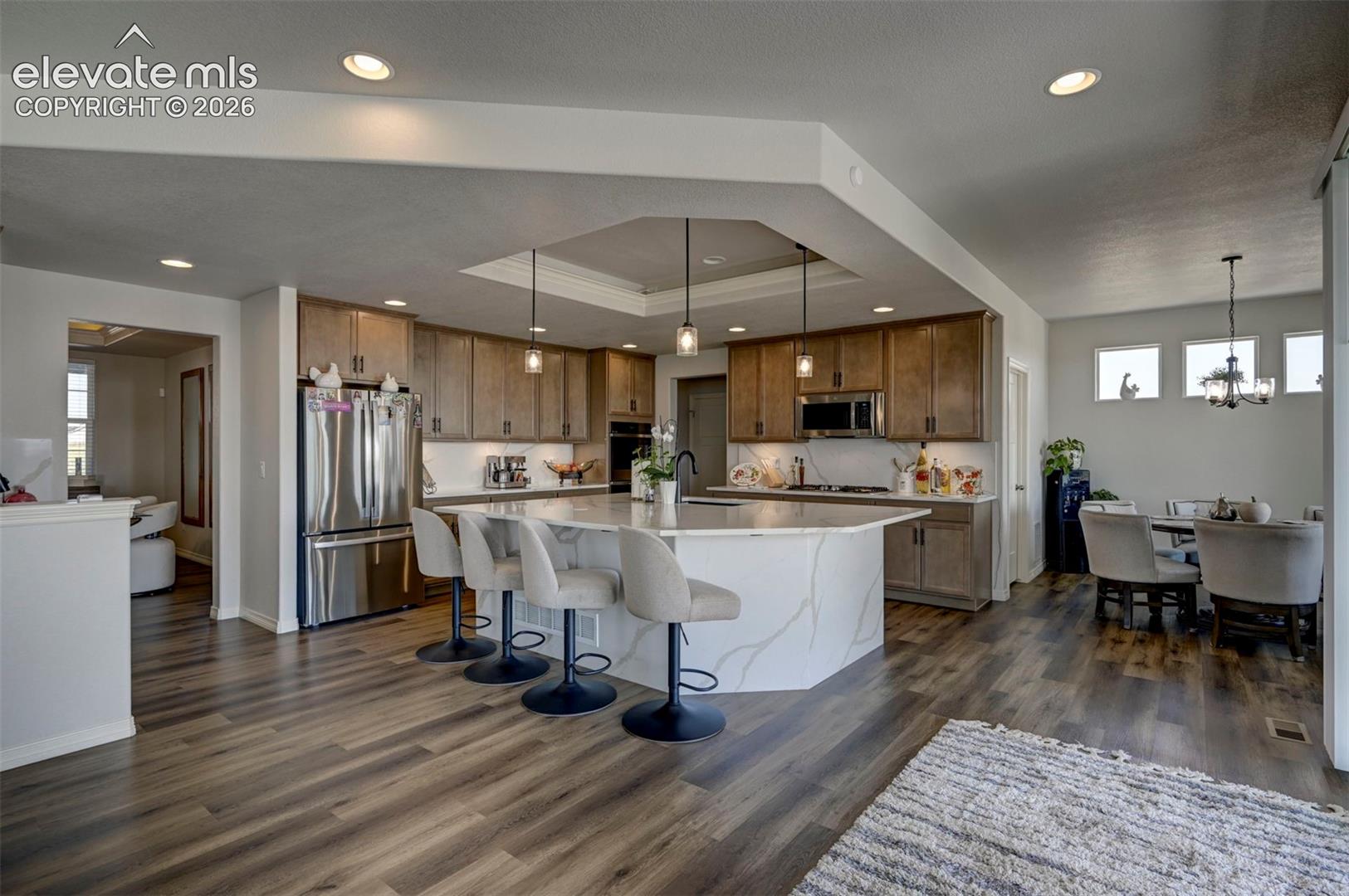 Contemporary kitchen featuring an oversized island with a waterfall-edge countertop, stainless steel appliances, wood-finish flooring, recessed lighting, and a tray ceiling