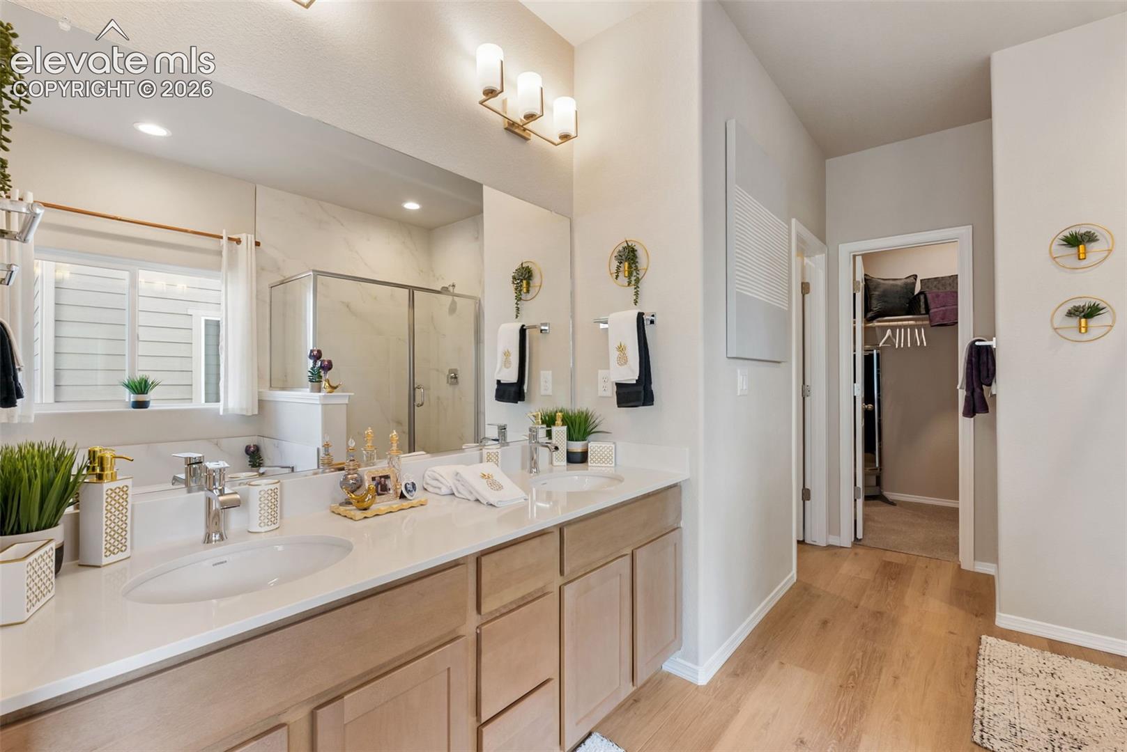 Dual-sink vanity with light-tone cabinetry and white countertops, large wall-mounted mirror, three-light polished finish fixture, walk-in shower with tile surround, and wood-finish flooring