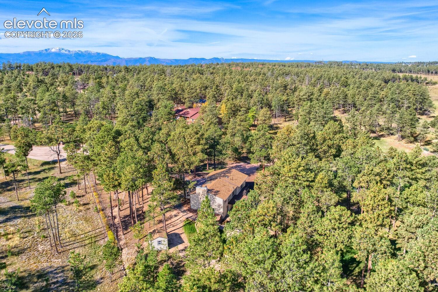 Expansive treed lot featuring a residence with a brown shingle roof, a secondary structure, and mountain scenery in the distance