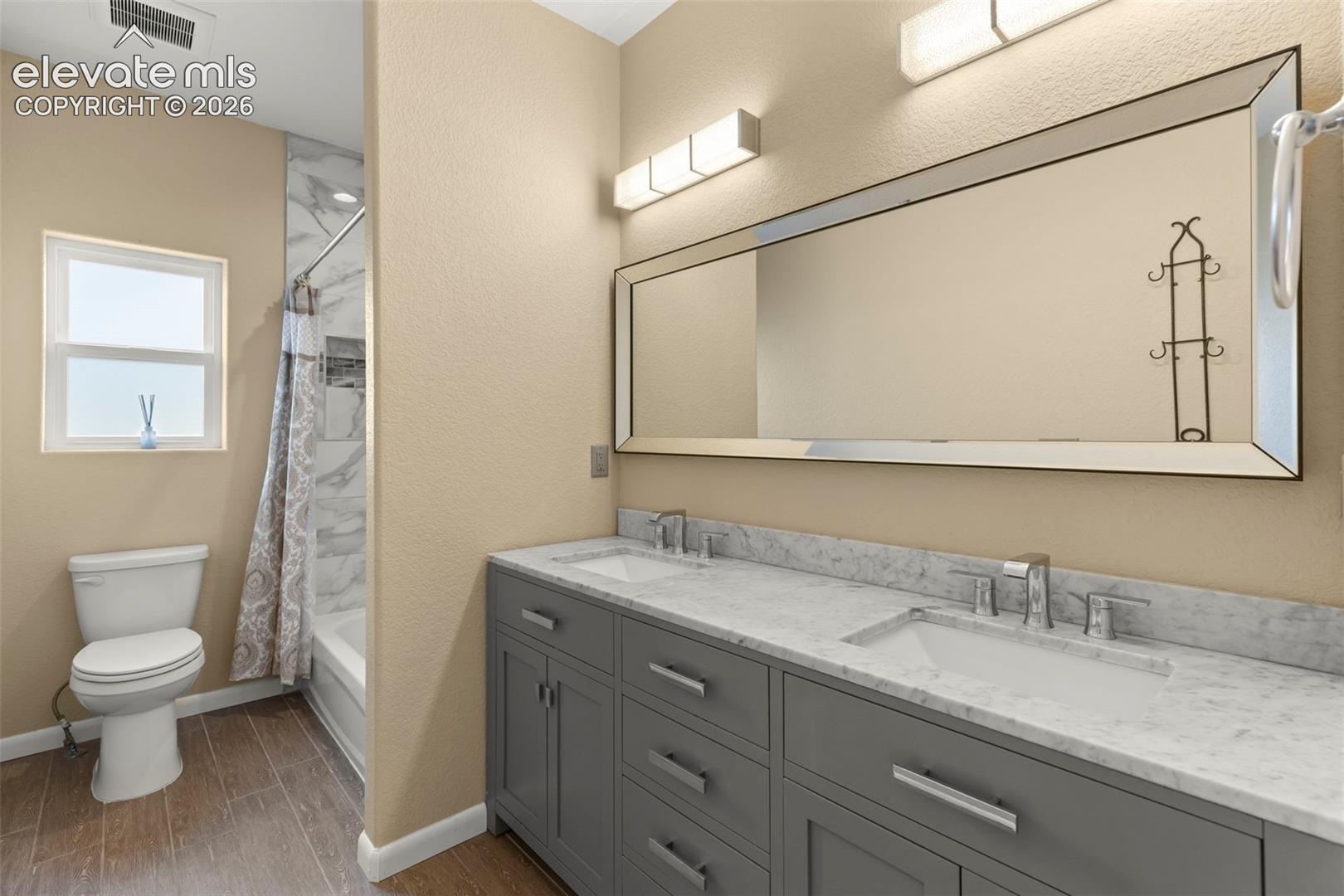 Double vanity featuring dark gray cabinetry, stone-finish countertops with dual integrated sinks, and polished chrome fixtures, complemented by wood-finish flooring and a tiled shower surround
