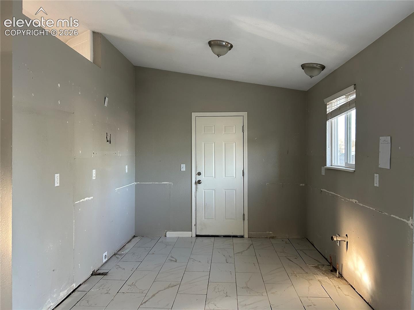 Interior space featuring large-format tile flooring with a neutral pattern, vaulted ceiling, dual flush-mount light fixtures, six-panel exterior door, and a single-hung window with built-in blinds
