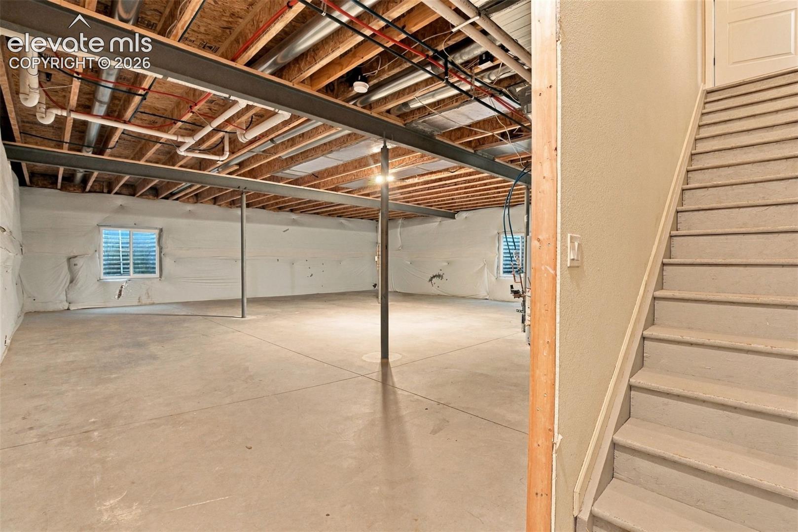 Unfinished basement space featuring exposed wood joists, structural steel support beams, concrete flooring, and two dual-pane windows