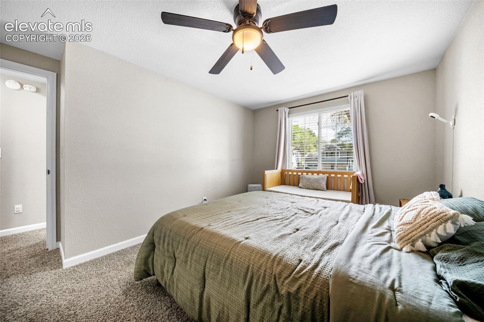 Spacious bedroom featuring neutral wall finishes, carpeted flooring, a ceiling fan with integrated lighting, and abundant natural light from dual windows with blinds and window seat storage