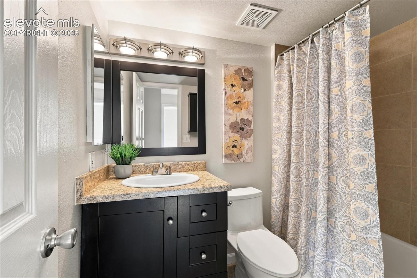 Bathroom featuring a dark-finish vanity with a stone-composite countertop, an integrated oval sink, a framed mirror with three-bulb vanity lighting, and tile-surround tub