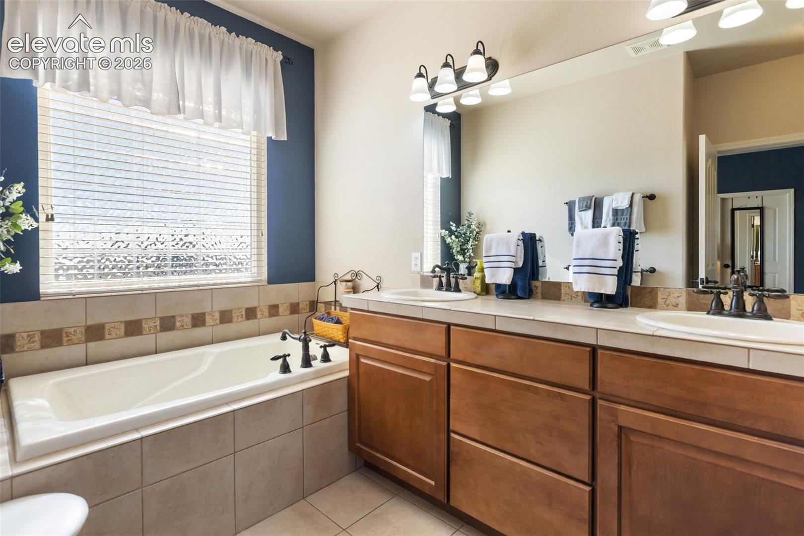 Primary Bathroom with Double vanity with tile countertop, drop-in soaking tub with tile surround, wood cabinetry, and multi-light wall fixtures