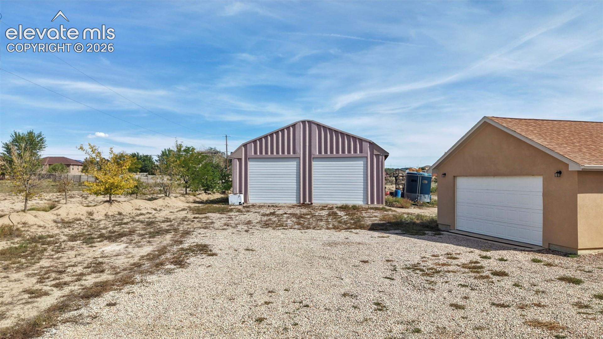 Detached metal outbuilding featuring dual roll-up garage doors, accompanied by a stucco-finish detached garage with a shingle roof and a gravel driveway