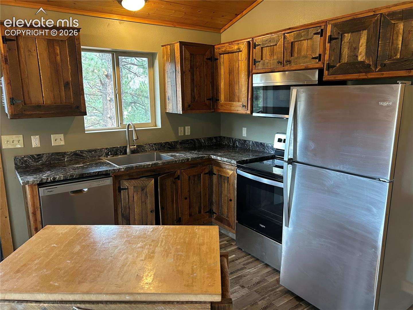 Kitchen featuring rustic wood cabinetry, dark stone-patterned countertops, stainless steel appliances, a wood-paneled vaulted ceiling, and wood-finish flooring