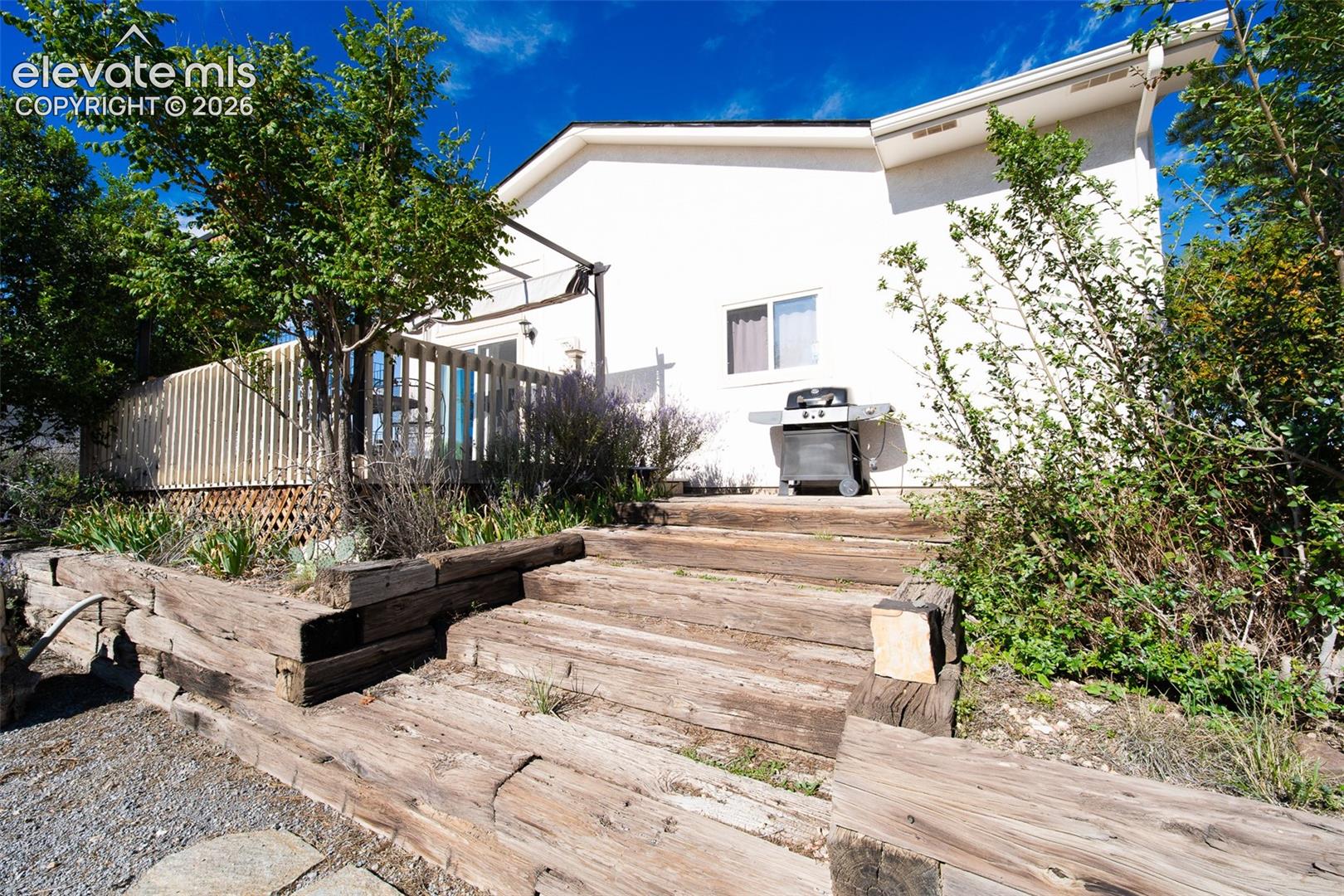 Rear exterior featuring a raised wood deck, tiered timber retaining walls, and mature landscaping