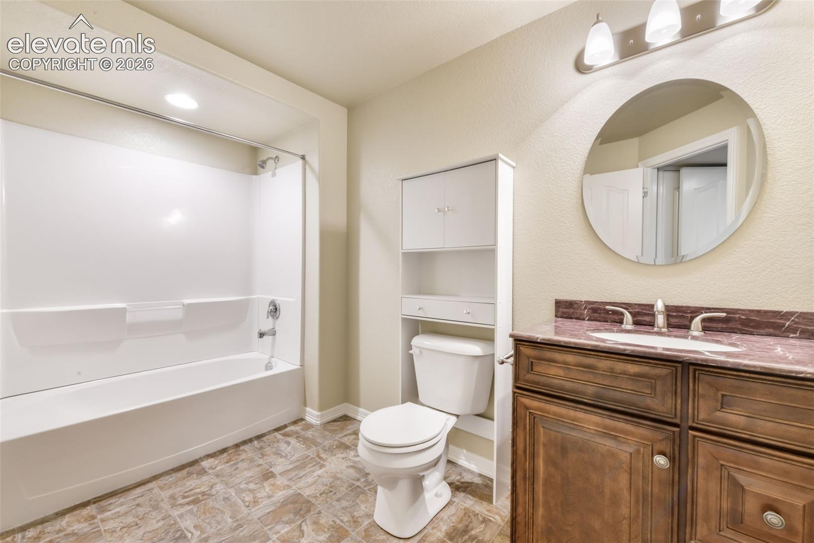 Bathroom featuring a tub/shower combination, dark wood-finish vanity with a single basin sink, integrated countertop, and a round wall-mounted mirror