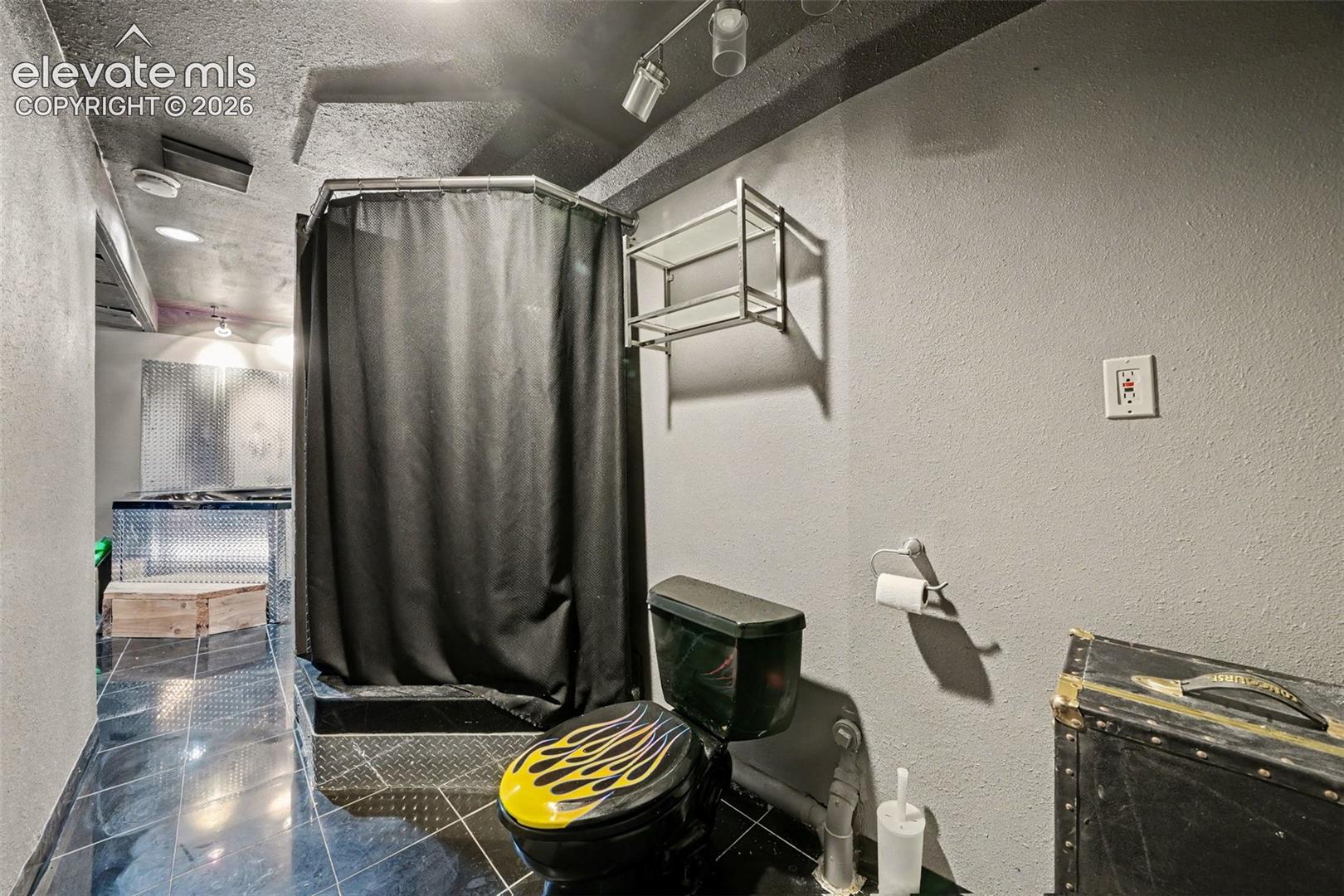 Bathroom featuring a corner shower with metallic diamond-plate detailing, wall-mounted glass shelving, track lighting, and dark tile flooring