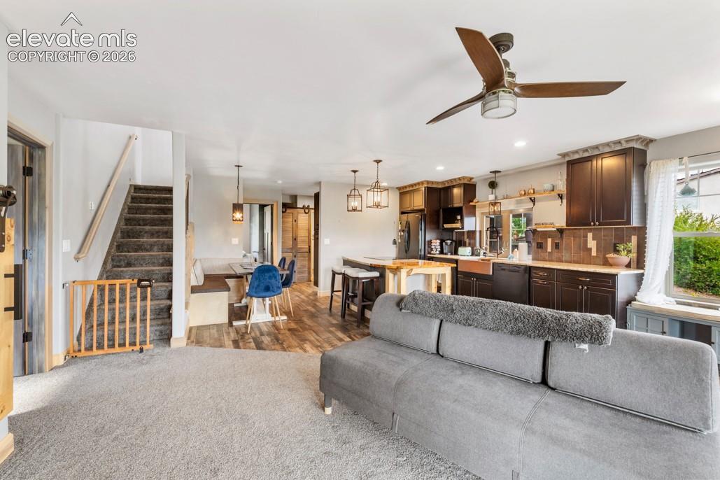 Open-concept living area with wood-finish flooring in the kitchen, dark wood cabinetry, a ceiling fan, and carpeted stairs