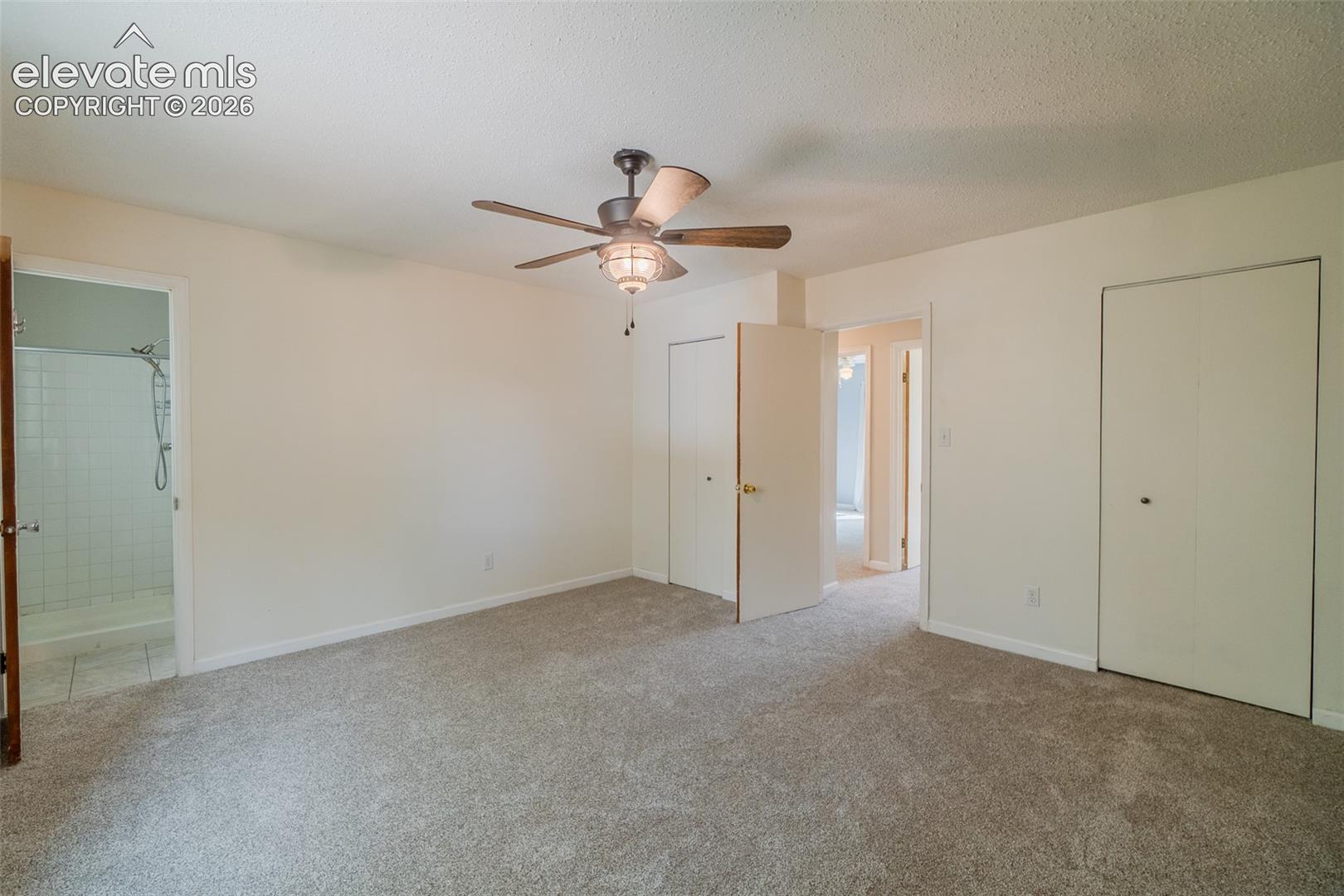Carpeted room featuring a ceiling fan with light fixture, two bi-fold door closets, and a private bath with a tiled shower