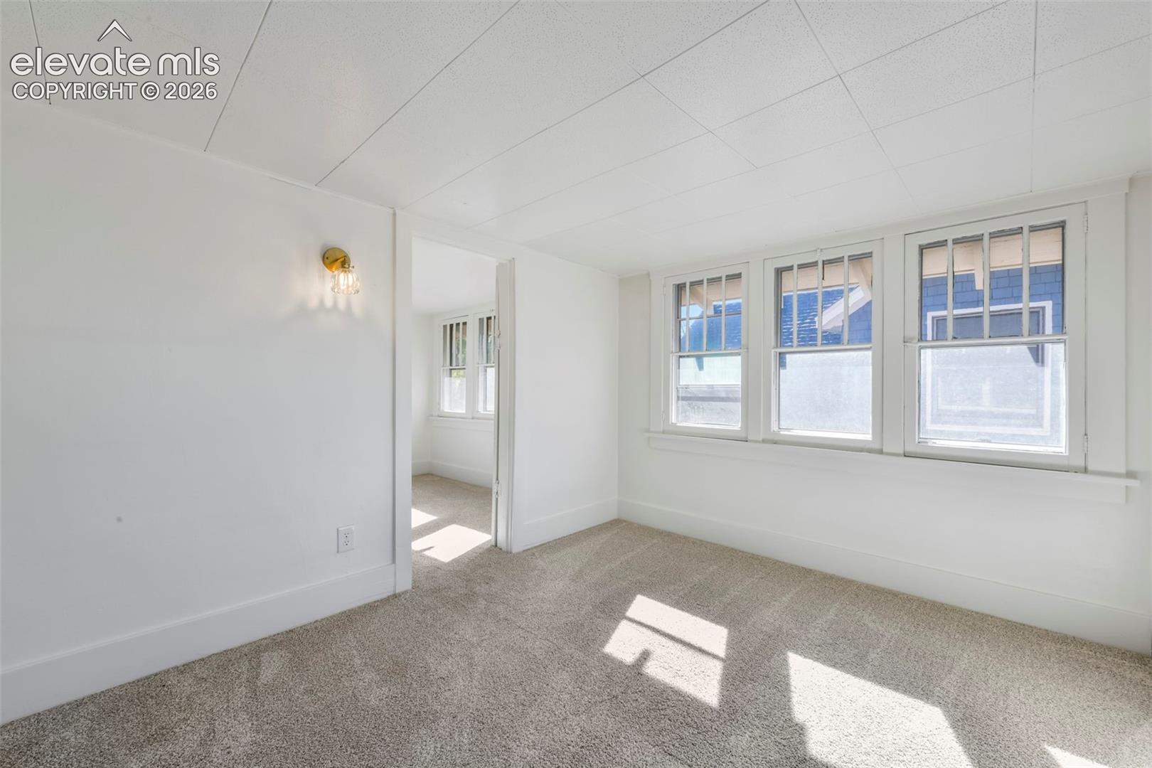 Interior space featuring wall-to-wall carpeting, three multi-pane windows with white trim, a decorative brass-finish wall sconce, and a clean, neutral color palette