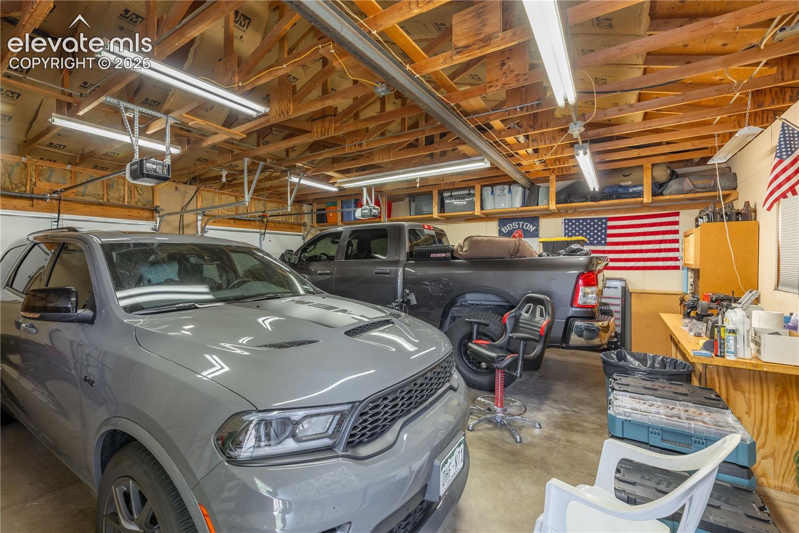 Attached garage featuring high ceilings with exposed wood rafters, linear fluorescent lighting, built-in overhead storage shelving, and a wood-surfaced workbench