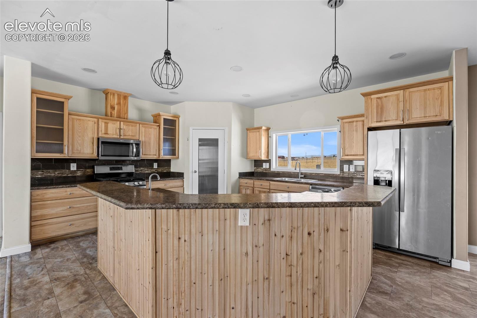 Kitchen featuring natural wood cabinetry, peninsula with vertical wood paneling, dark stone-finish countertops, stainless steel appliances, and dual metal-cage pendant lights