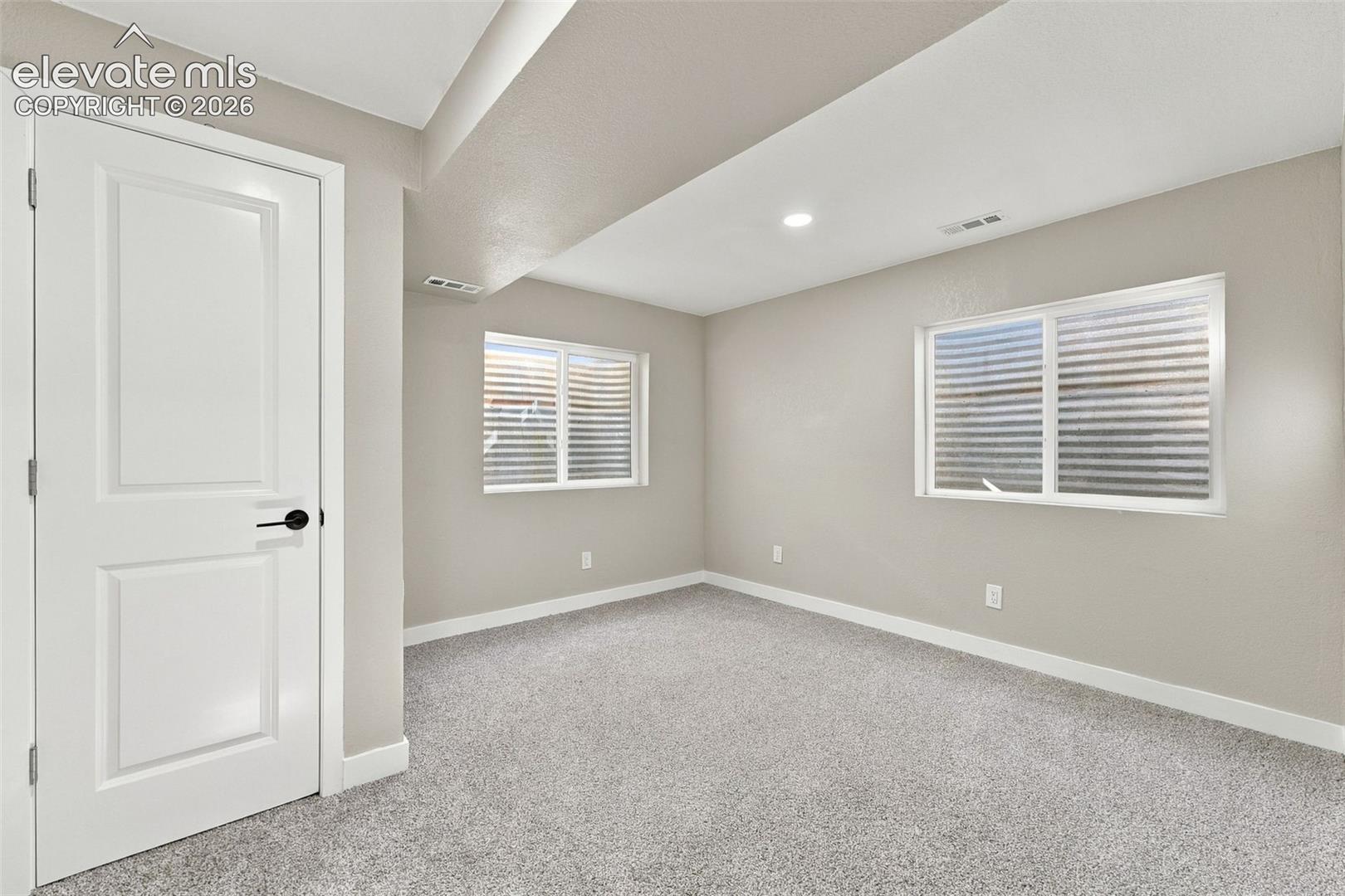 Neutral-toned interior space featuring wall-to-wall carpeting, two egress windows with corrugated window wells, recessed ceiling lighting, and a white interior door with matte black hardware