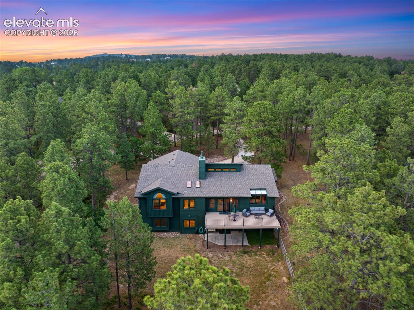 Wooded lot featuring a dark green exterior, multi-level roofline, ample windows, and an elevated outdoor deck