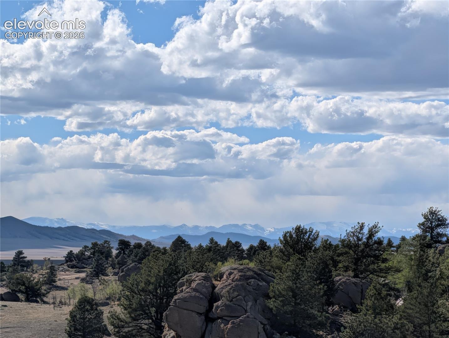 Expansive natural landscape featuring a prominent rock formation, numerous evergreen trees, distant snow-capped mountains, and a vast sky with cumulus clouds