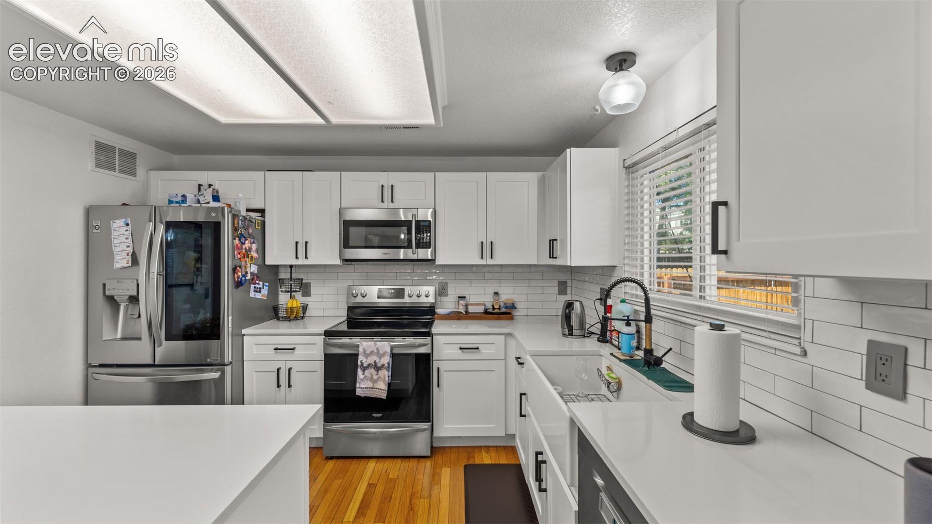 White shaker-style cabinetry with black hardware, subway tile backsplash, white countertops, built-in stainless steel appliances, and wood-finish flooring