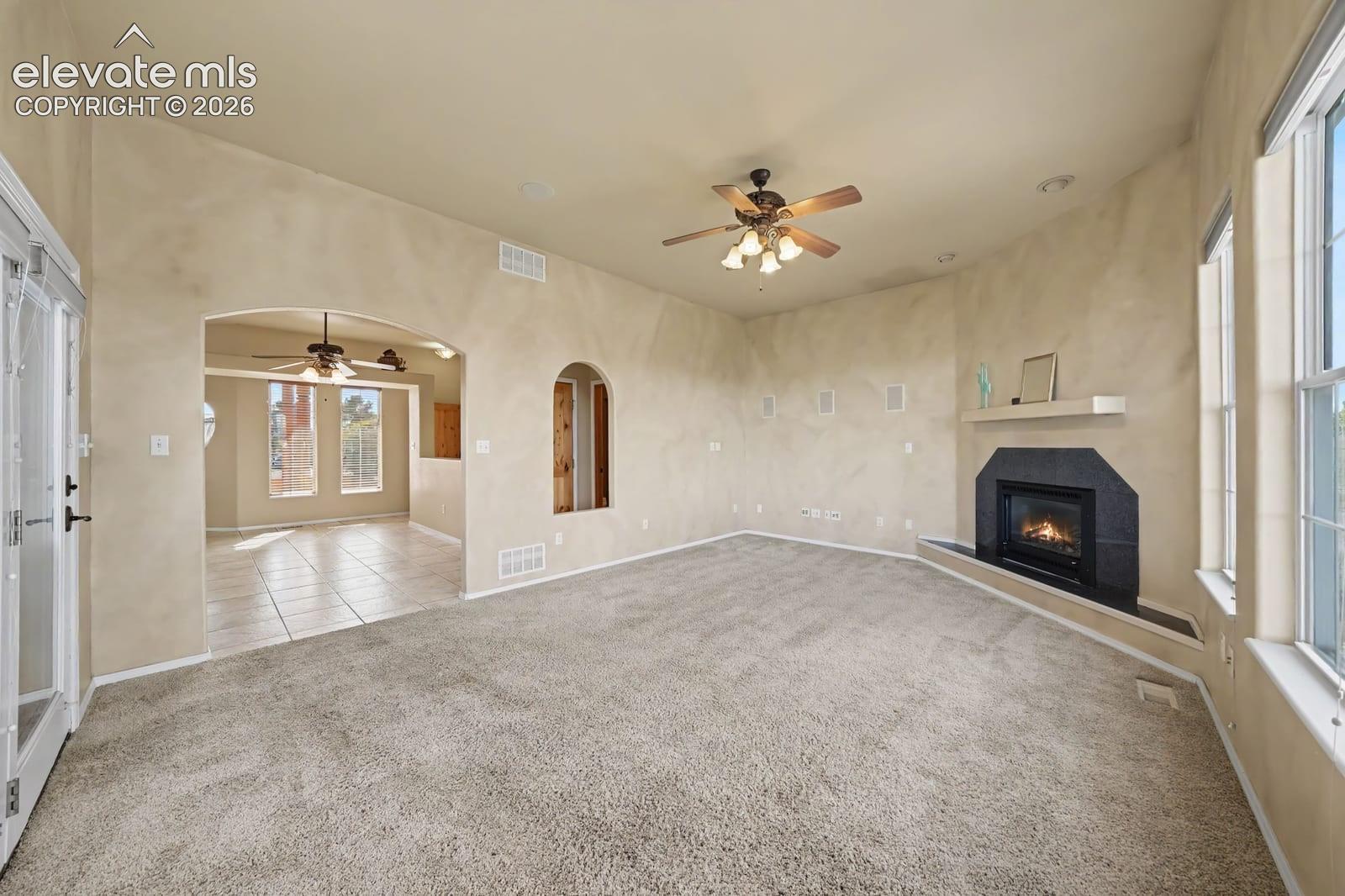 Living area featuring textured wall finishes, a fireplace with a stone-surround hearth and mantel, arched architectural openings, multi-pane windows, and a ceiling fan with integrated lighting