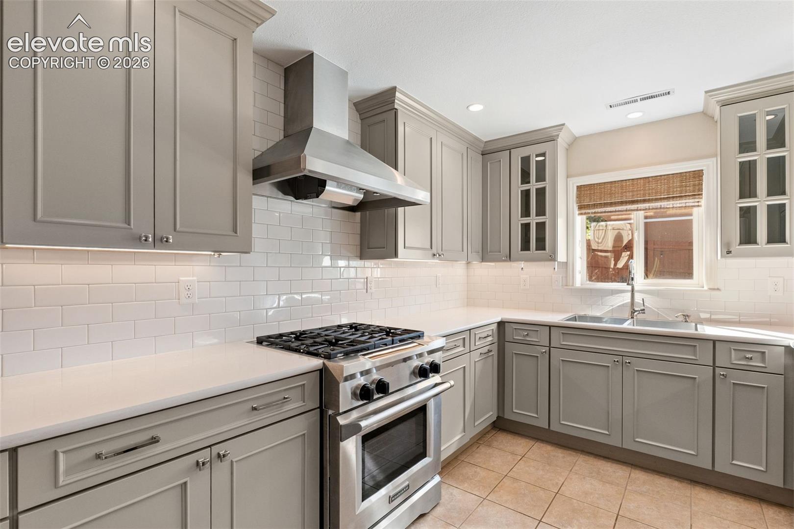 Shaker-style cabinetry, white subway tile backsplash, stainless steel gas range with overhead vent hood, dual-basin stainless steel sink, and solid-surface countertops