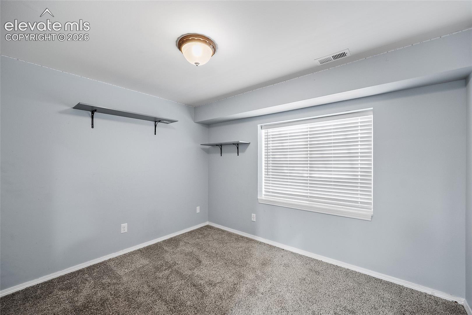 Recessed window with blinds, ceiling-mounted light fixture, wall-mounted shelving, and gray carpeting
