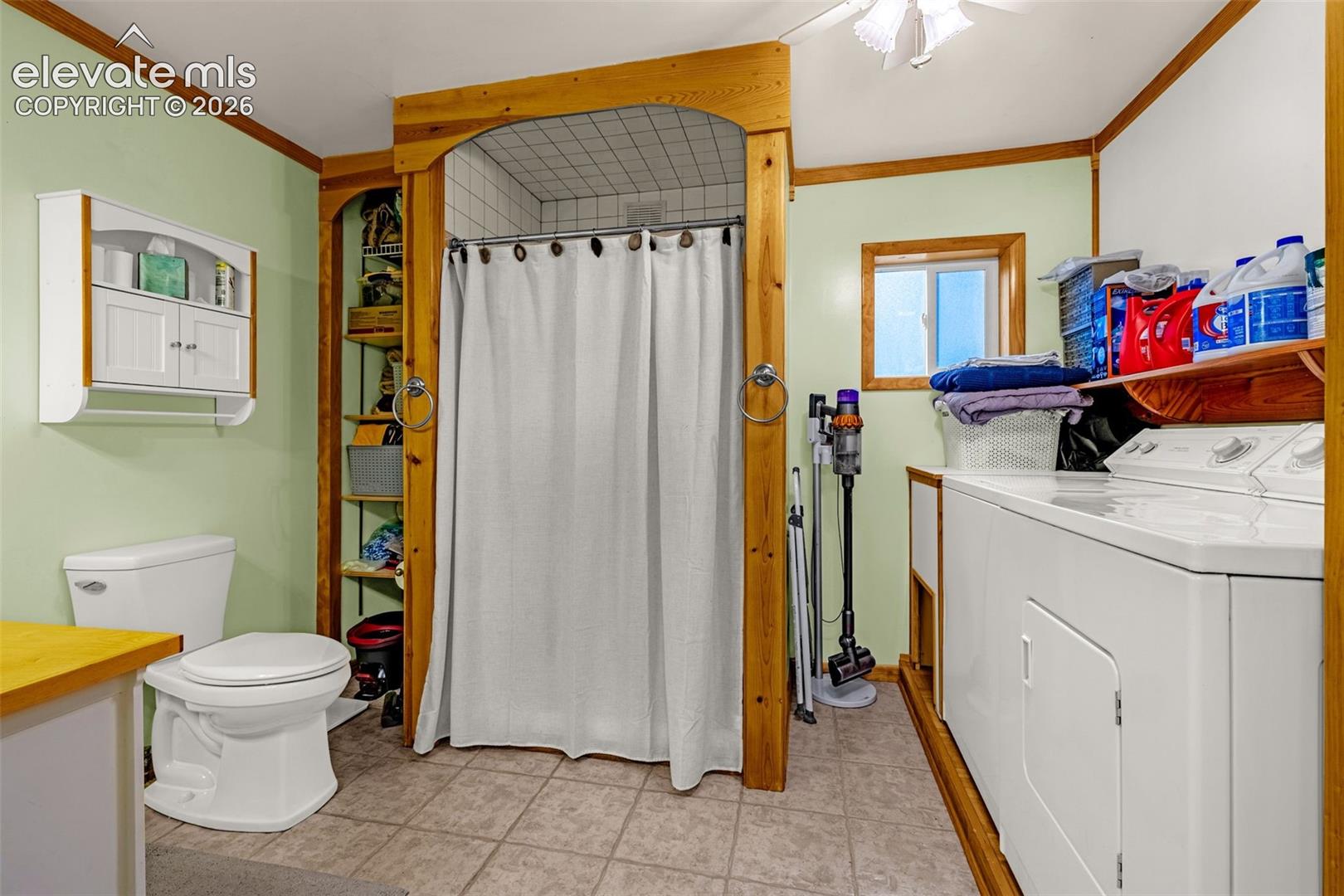 Bathroom featuring ceramic tile flooring, a shower stall enclosed by wood-finished trim, built-in shelving, white wall-mounted cabinetry, and dedicated laundry hookups