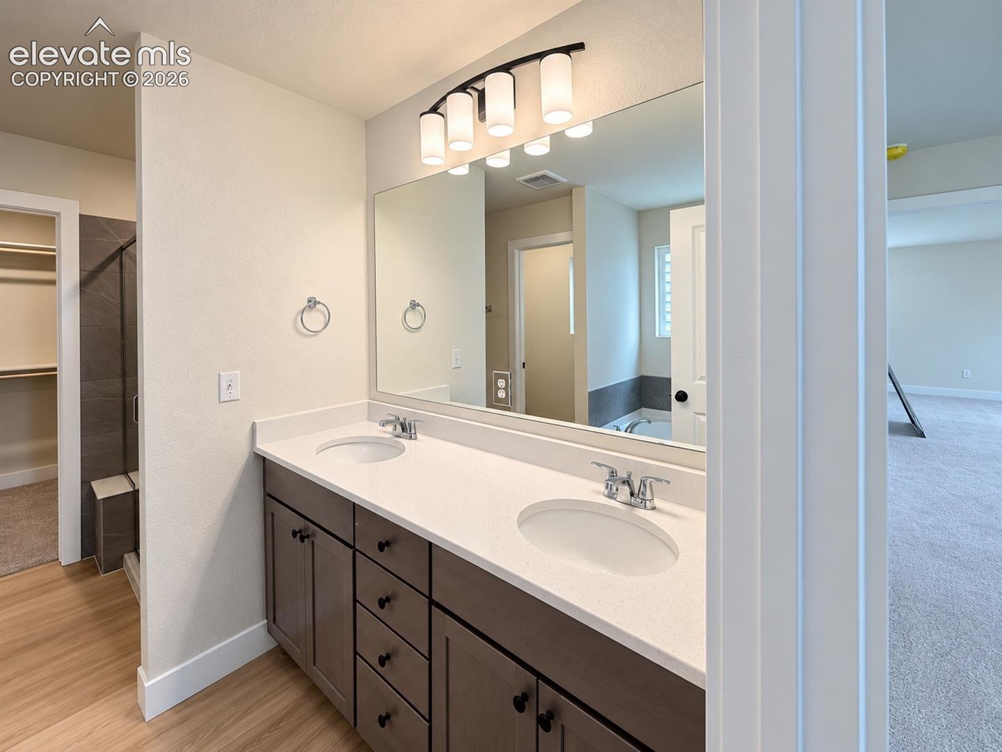 Dual-sink vanity featuring stone-surfaced countertops, custom-finish cabinetry, polished chrome fixtures, and a large plate-glass mirror with multi-bulb overhead lighting