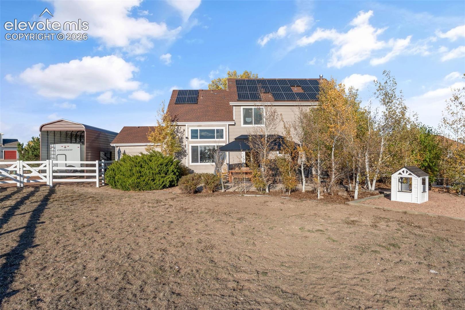 Two-story residence featuring solar panel arrays, a detached metal-frame carport, and a white rail fence enclosing a leveled lot