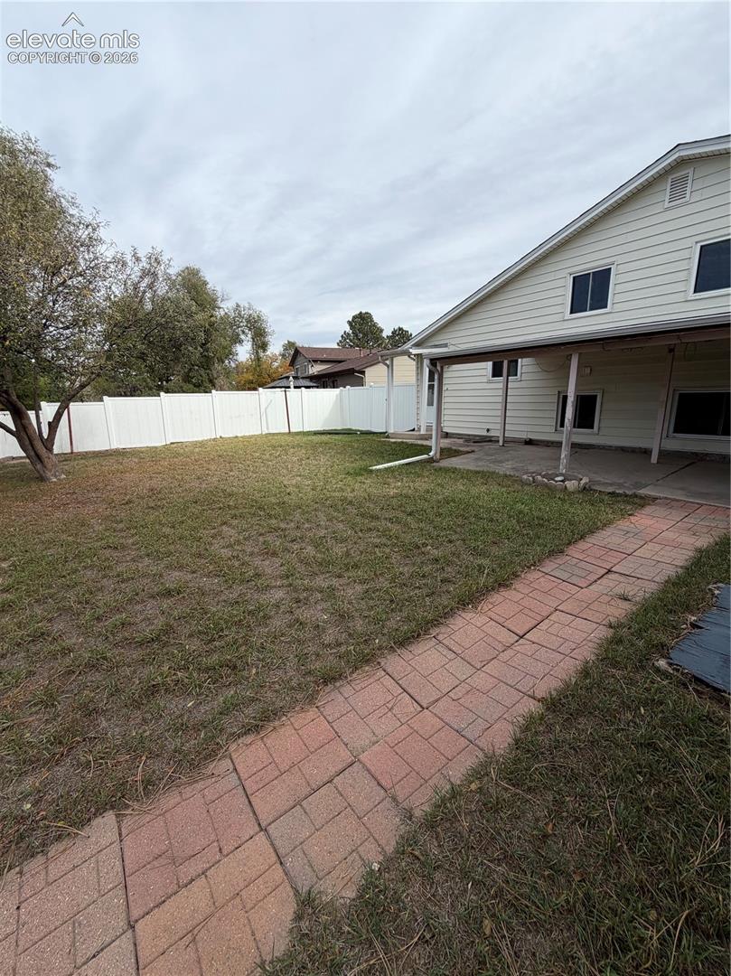 Backyard featuring a paver walkway, covered concrete patio, white vinyl privacy fencing, and mature trees