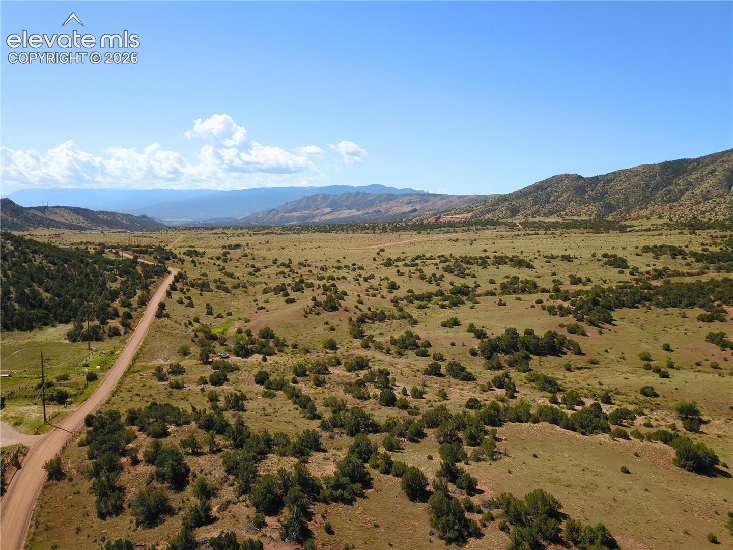 Expansive open land featuring a dirt road, scattered green vegetation, and distant mountains under a clear sky
