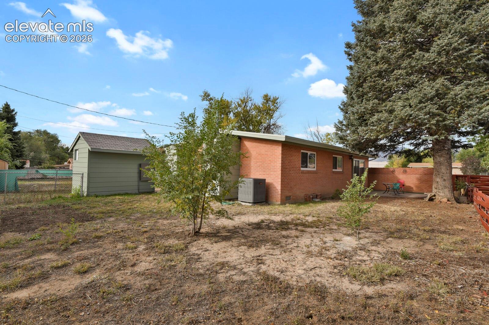 Rear exterior featuring red brick siding, a detached storage structure, concrete patio, and mature evergreen tree. Plenty of room for activities or a Garden Area.  