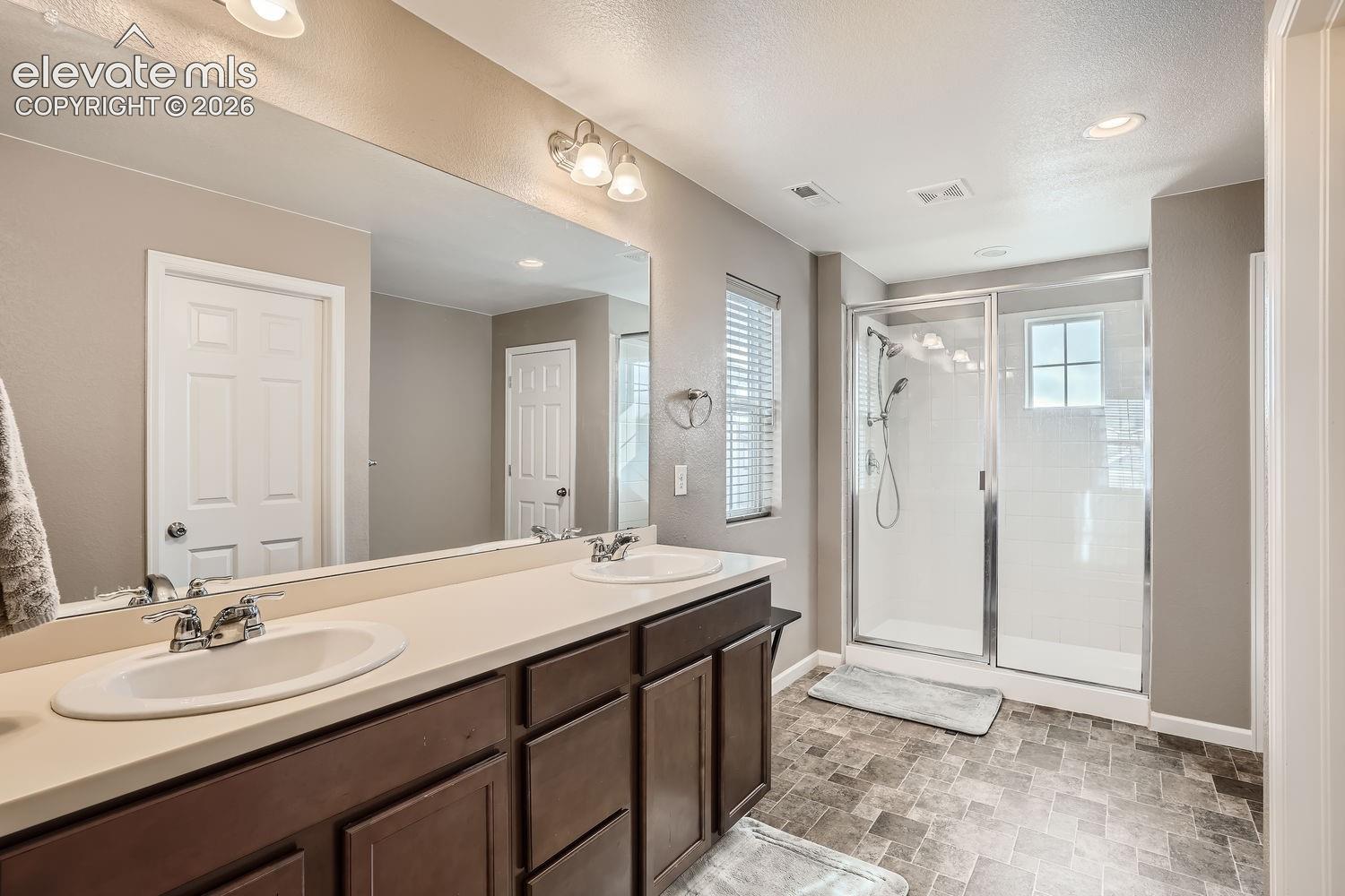 Dual-sink vanity featuring a solid surface countertop, dark wood cabinetry, a full-width mirror, and a walk-in shower with a glass enclosure and tile surround