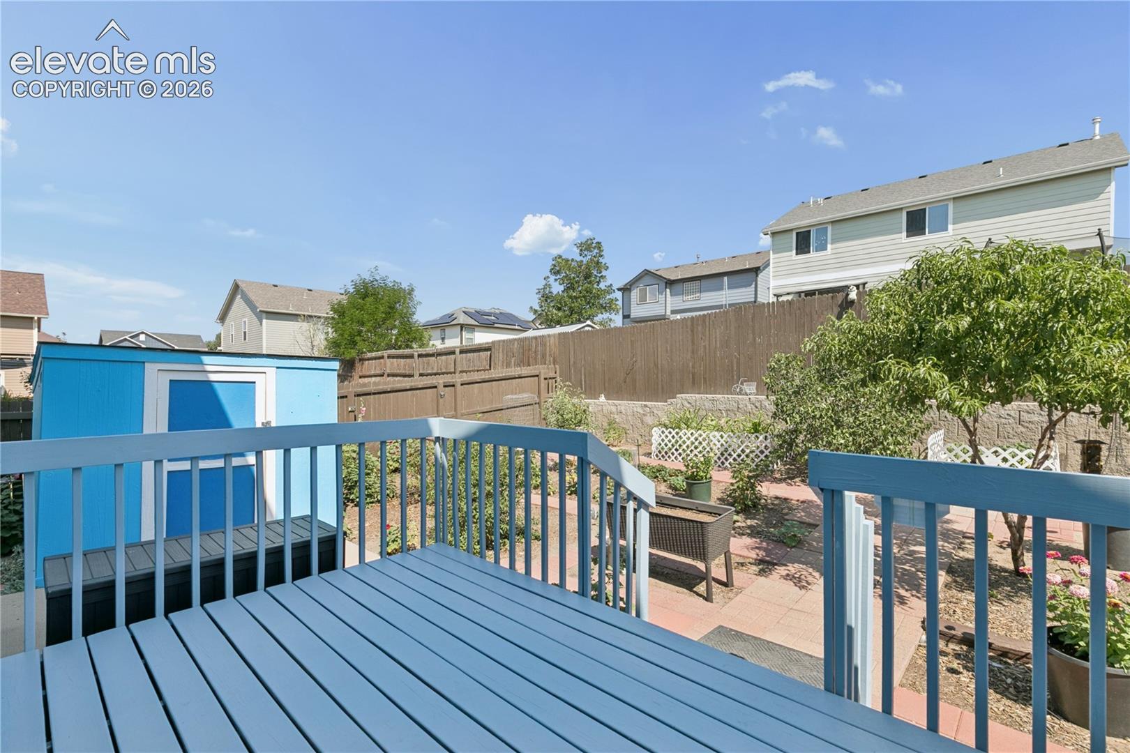 Spacious wooden deck with blue paint, leading to a backyard featuring a blue storage shed, privacy fencing, and mature landscaping