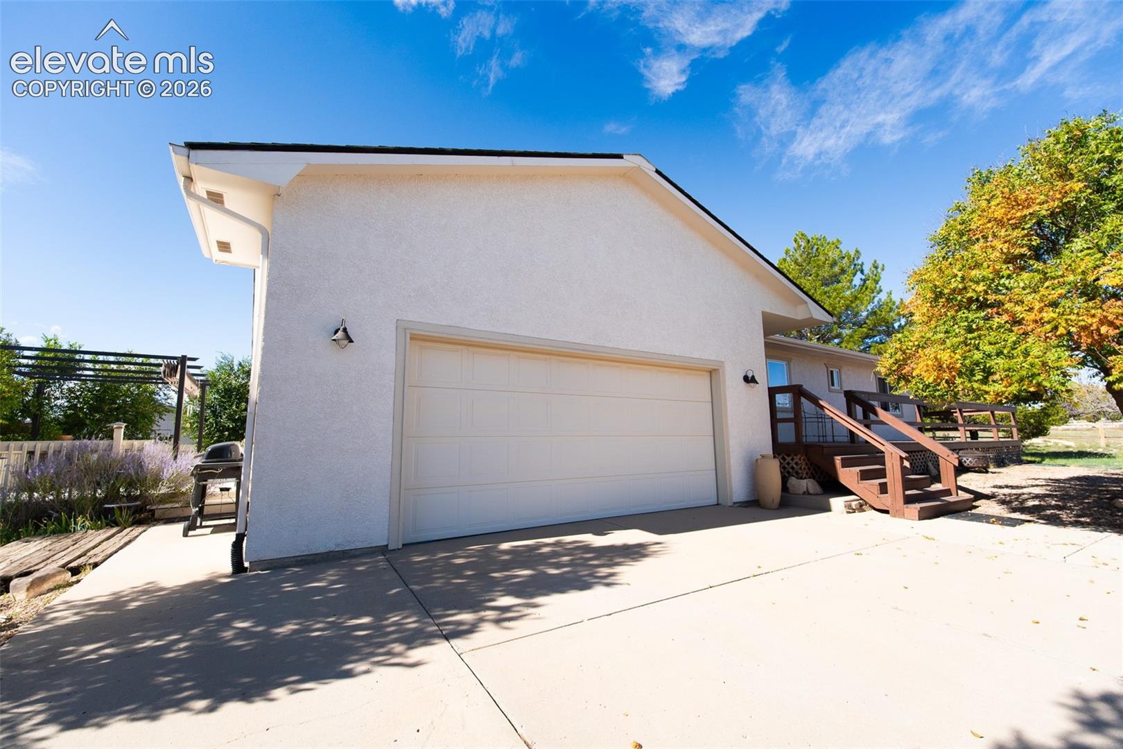 Stucco exterior featuring a two-car garage, concrete driveway, wood-framed deck, and metal pergola
