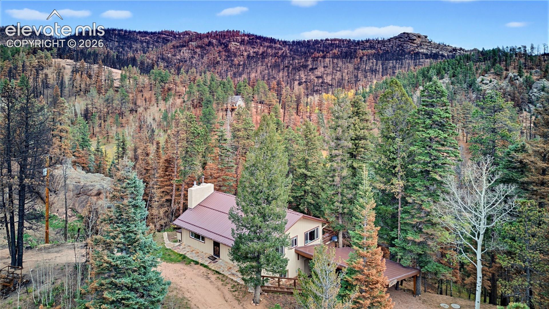 Mountain residence featuring a metal roof, exterior chimney, and tiered wooden decking, set against a backdrop of mountainous terrain and mature pine forest