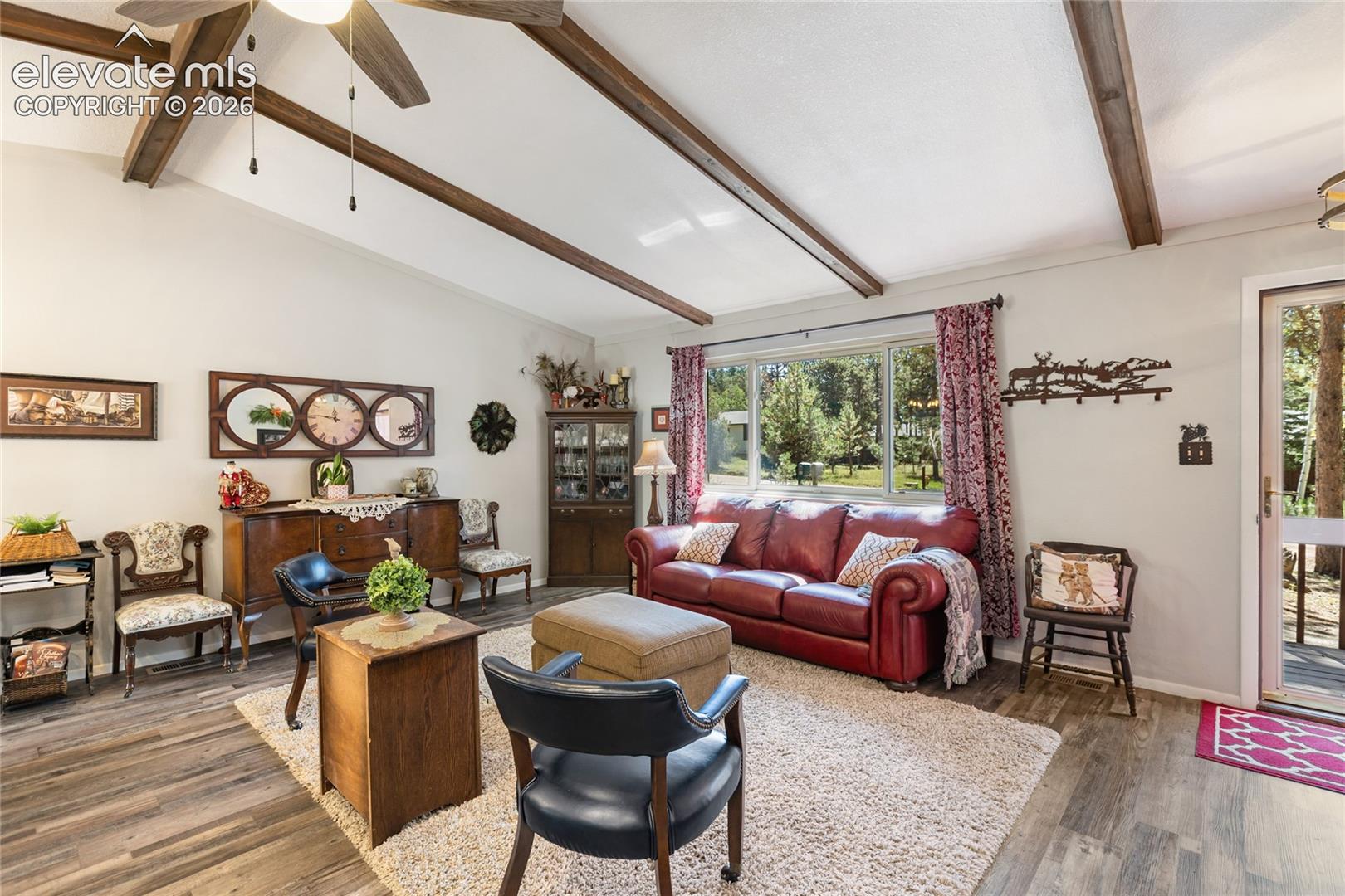 Vaulted ceiling with exposed wood beams, wood-finish flooring, large window with outdoor scenery, and a glass-paneled exterior door