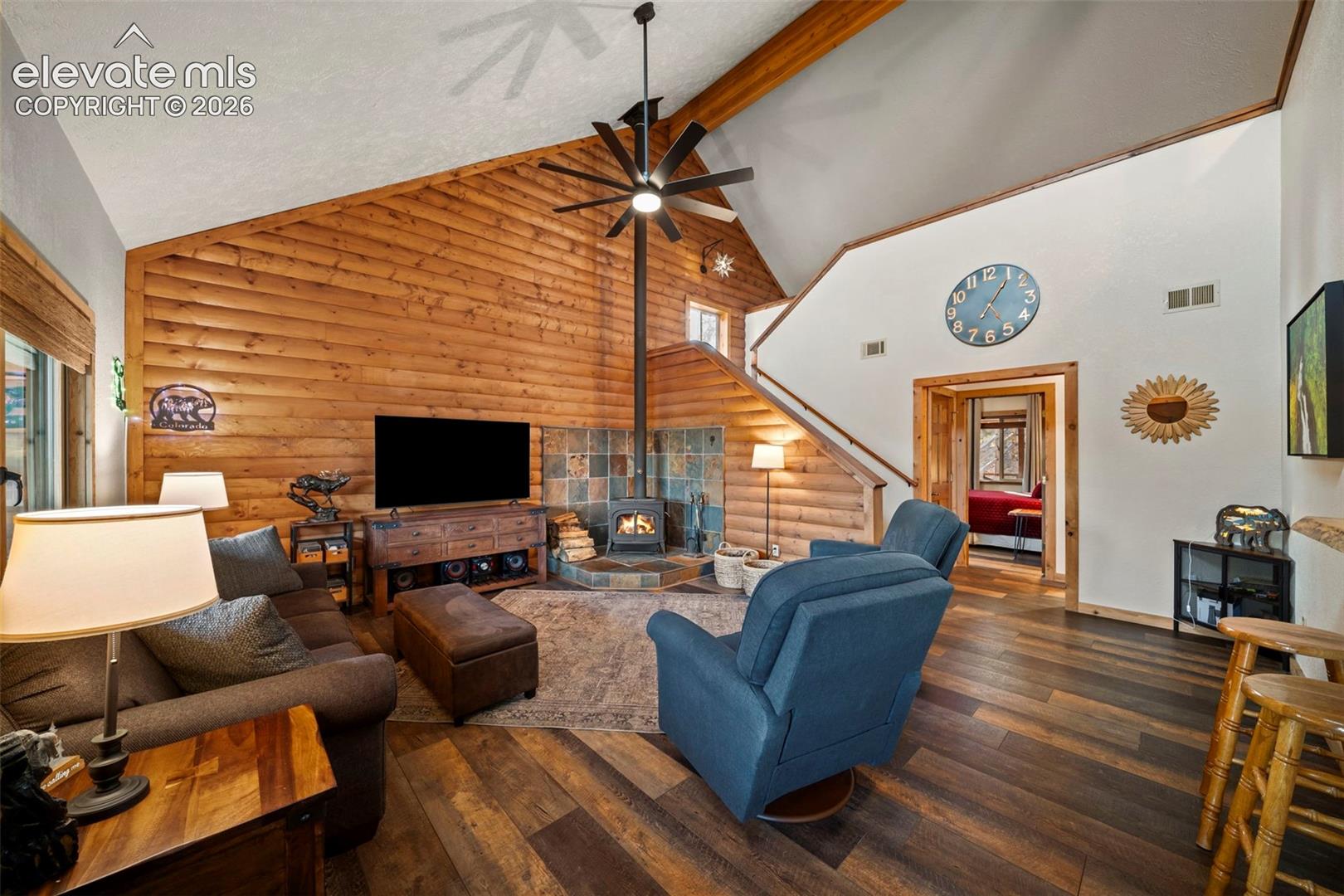 Vaulted ceilings featuring exposed timber beams, wood-plank wall paneling, tile-surround corner stove, and wide-plank wood-finish flooring