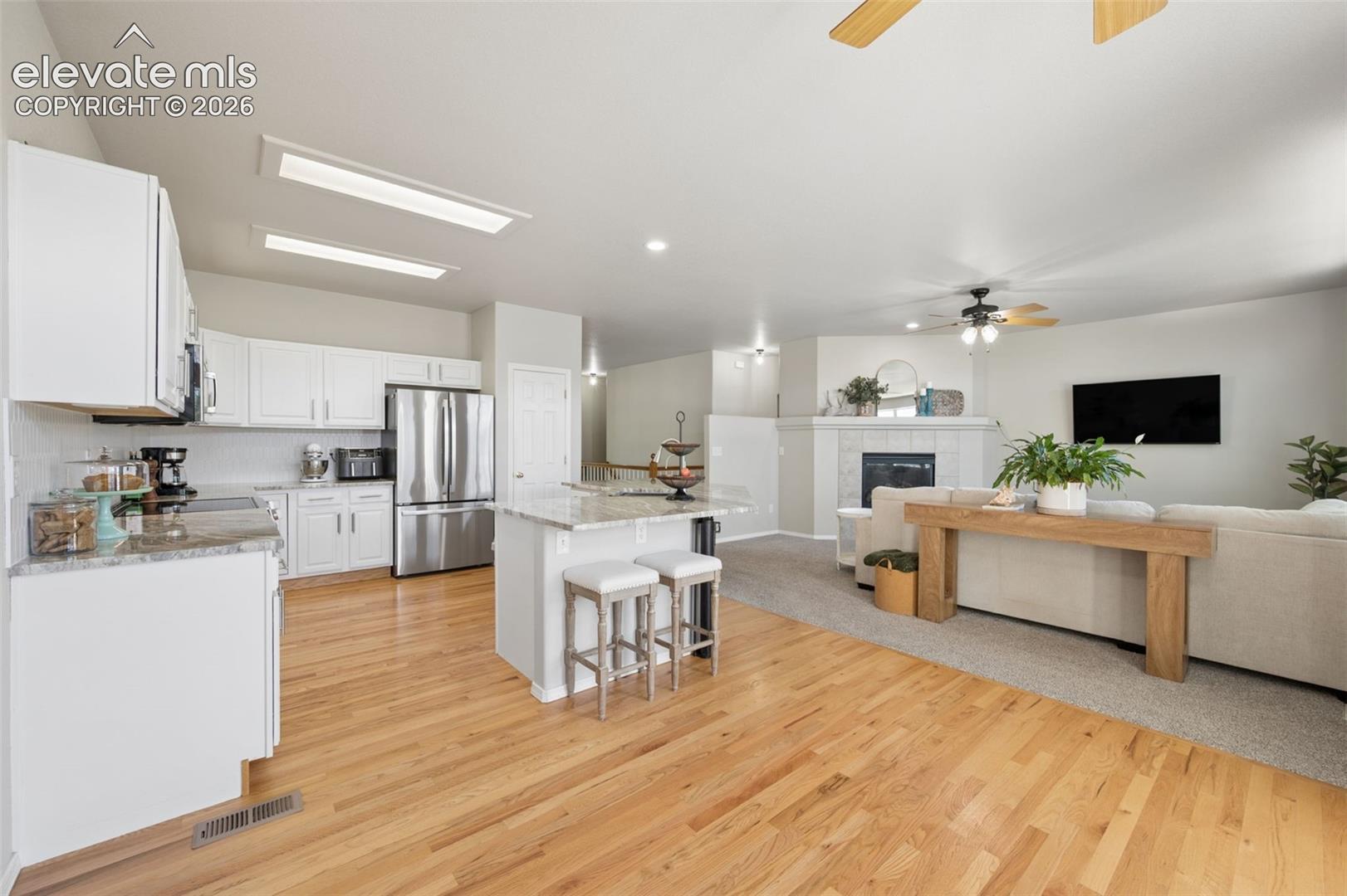 Kitchen featuring white shaker-style cabinetry, stone-finish countertops, an island, wood-finish flooring, and a fireplace with a tiled surround
